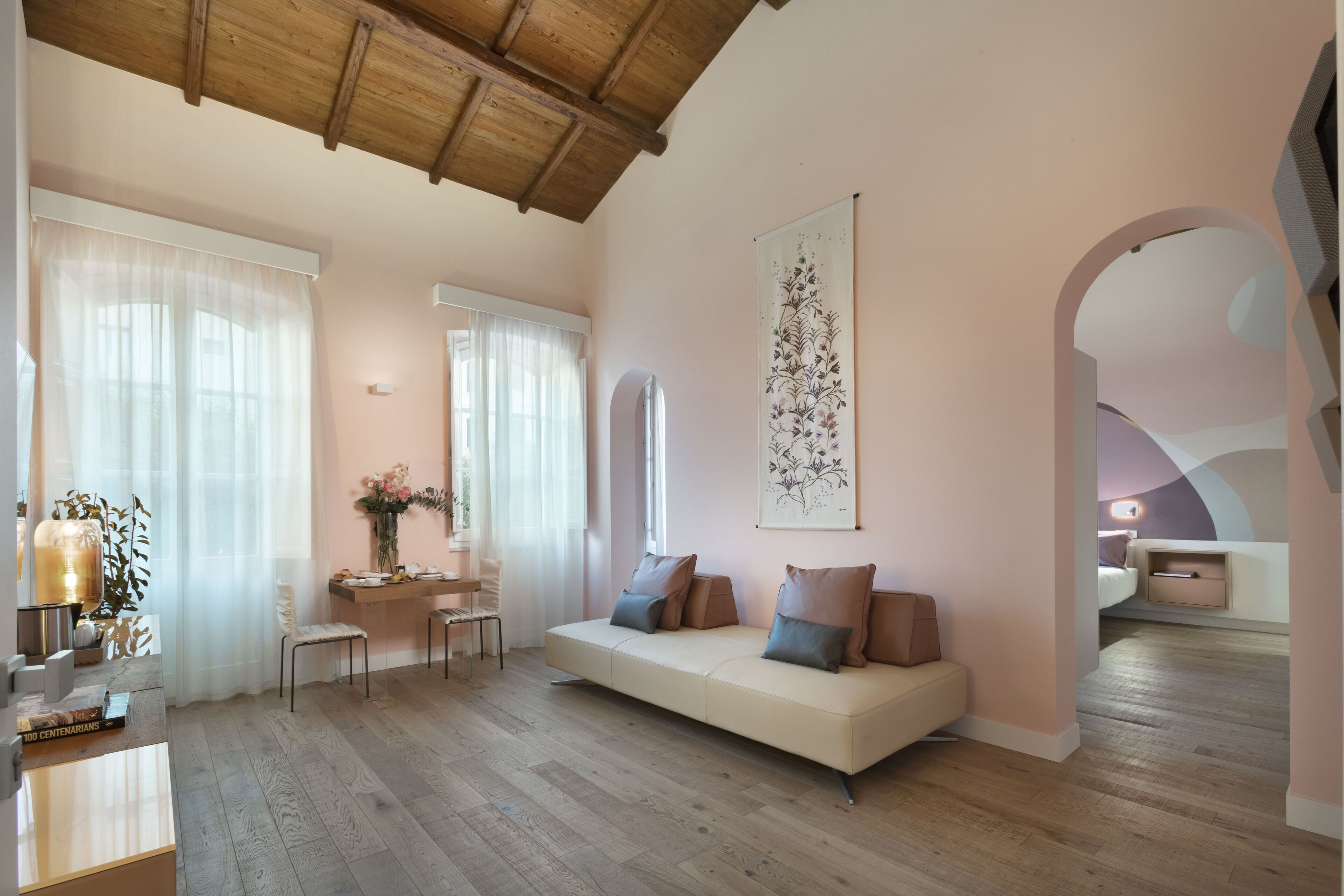 A modern living room with a wooden ceiling, a sofa with cushions, a dining area with a table and chairs, and a large window with sheer curtains.