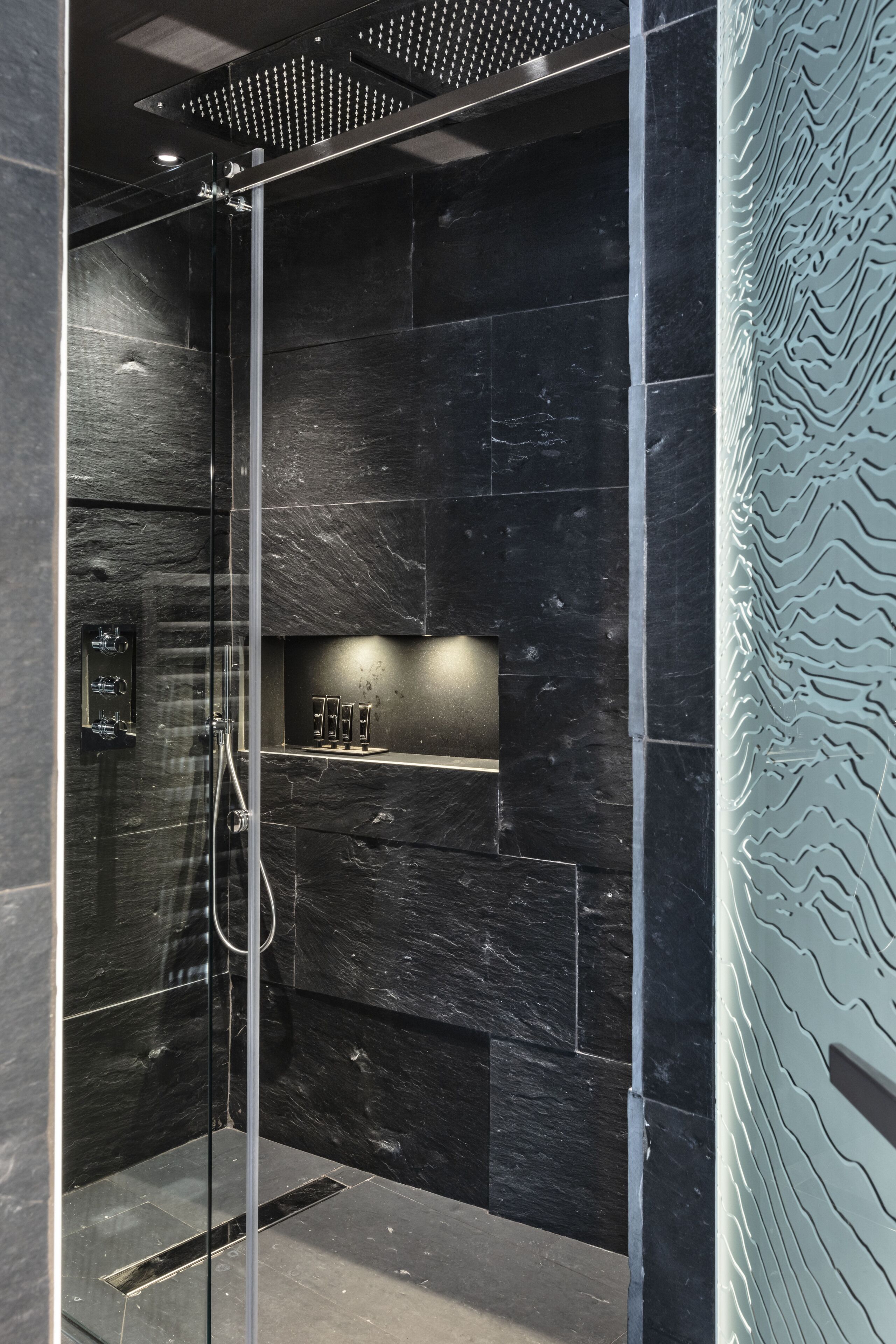 A modern shower with dark marble walls, a glass door, and a shelf with toiletries.