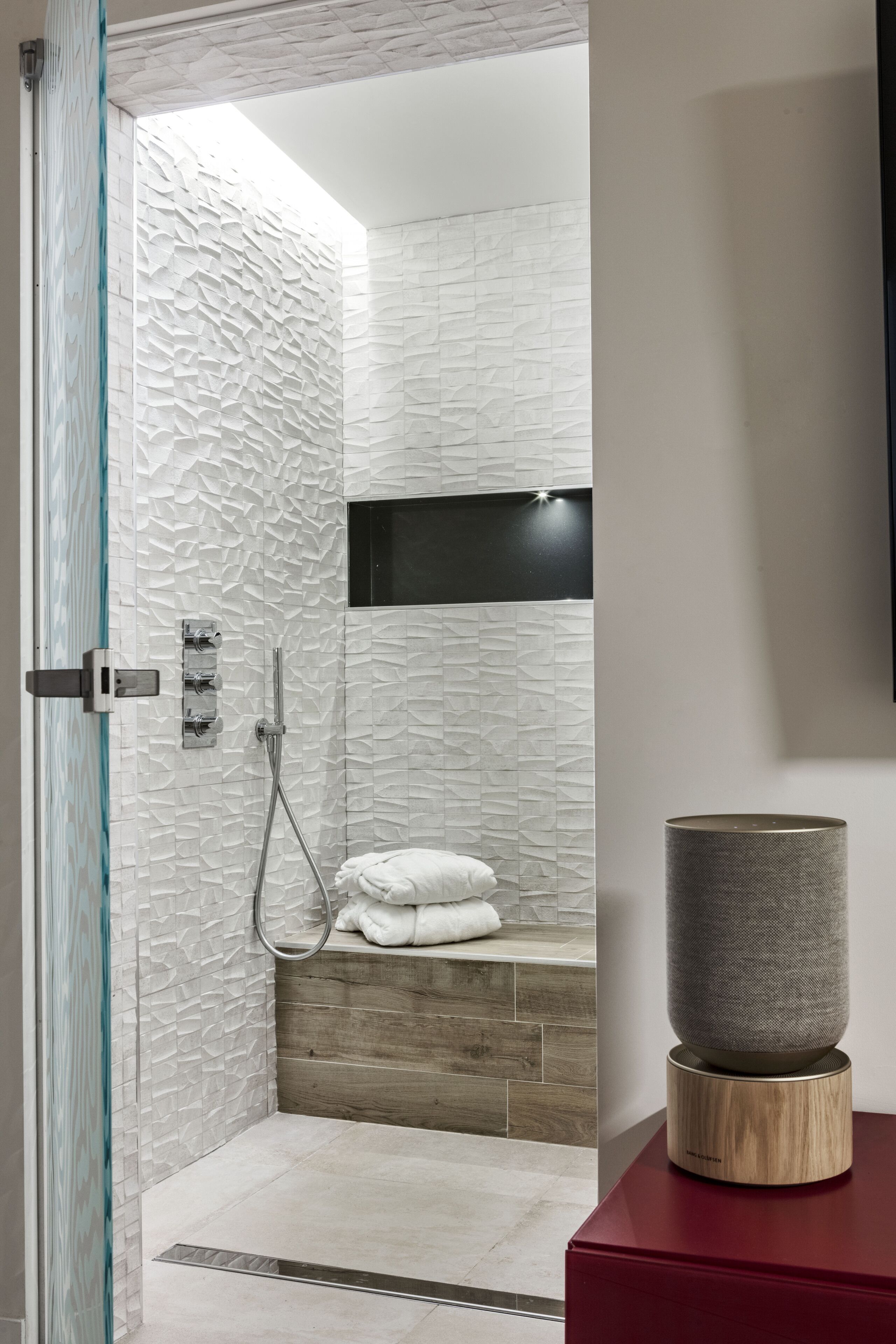 Een moderne badkamer met gestructureerde witte wanden, een houten bankje, een douche met glazen deur en een rode wastafel met een speaker.