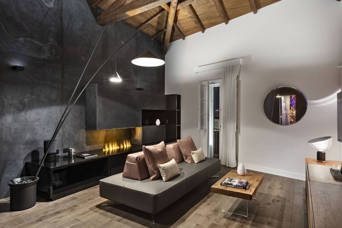 Ein modernes Wohnzimmer mit dunkler Steinwand, Holzboden und einer Couch mit Kissen, einem Couchtisch und einem runden Spiegel an der Wand.