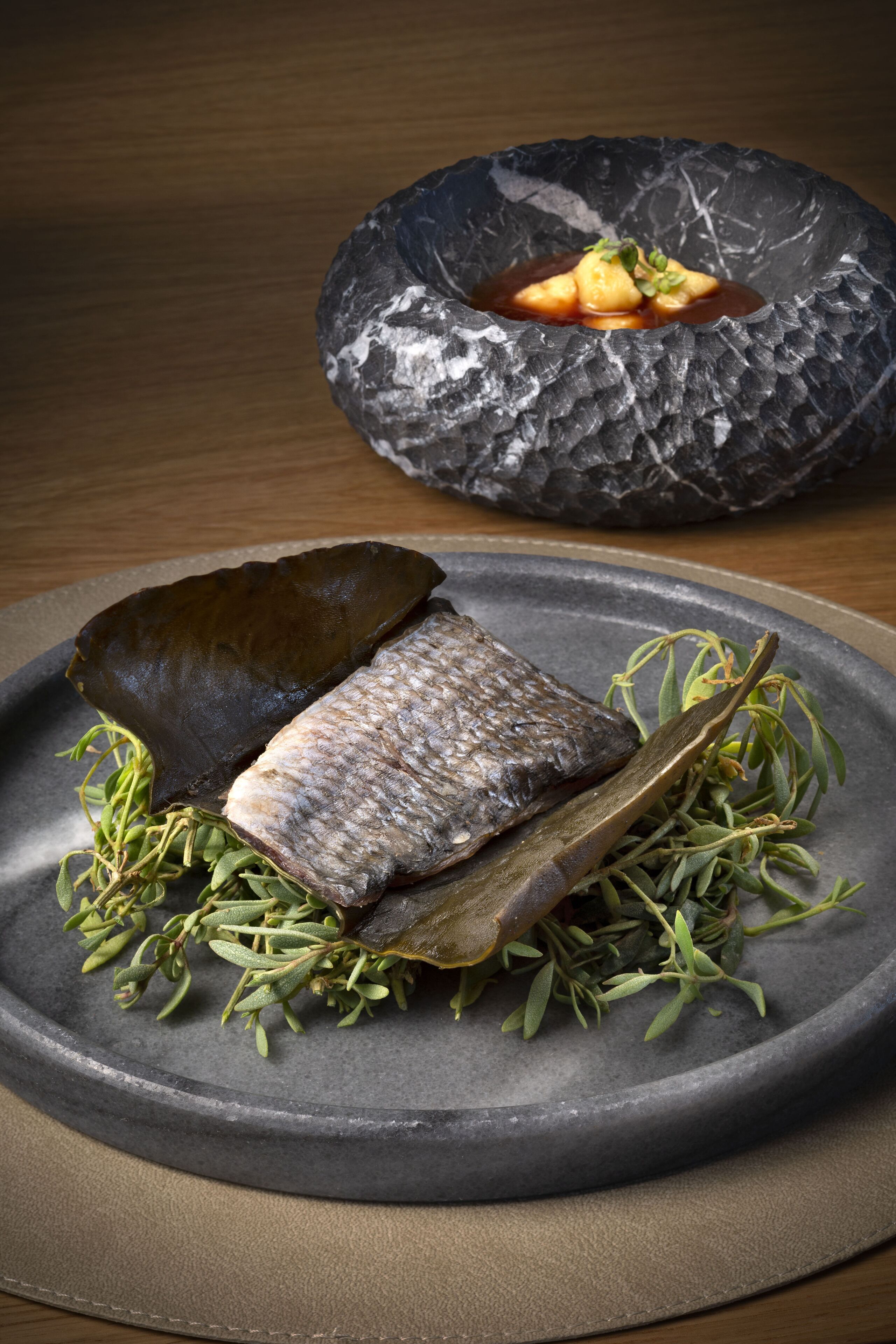 A stone plate with a fish, herbs, and a dark bowl with a sauce garnish.