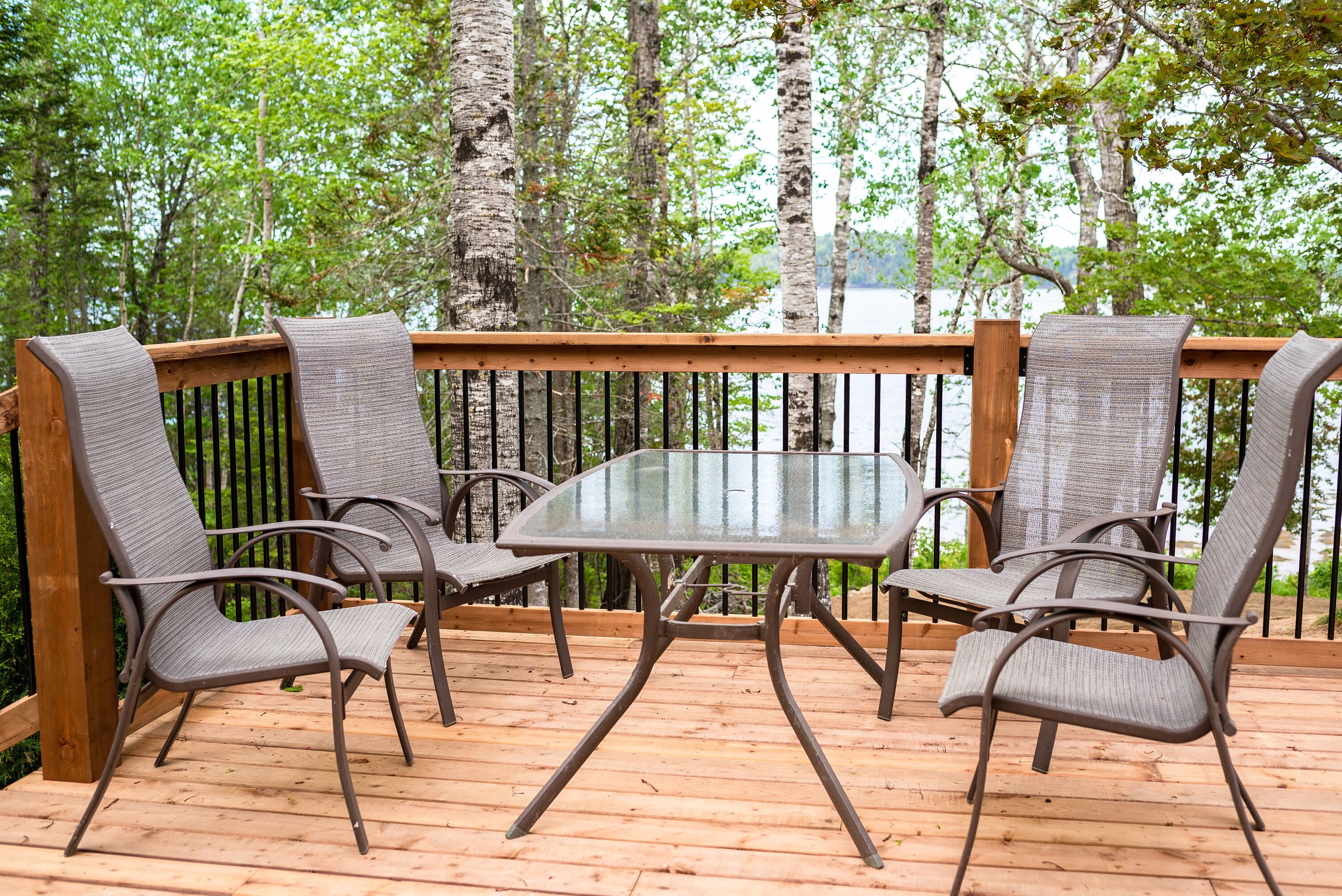 A wooden deck with a glass table and four chairs, surrounded by a wooden railing and trees.