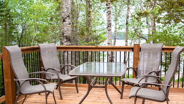 A wooden deck with a glass table and four chairs, surrounded by a wooden railing and trees. - Woodhaven Country Lodge (Oak Bay)