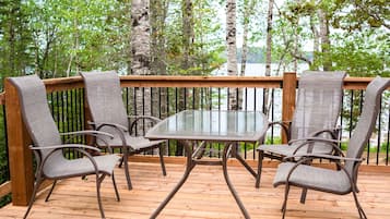 A wooden deck with a glass table and four chairs, surrounded by a wooden railing and trees.