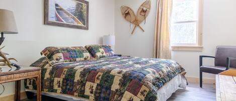 Ein Schlafzimmer mit einem großen Bett, einem Nachttisch, einem Sessel, einem Fenster mit Vorhängen und einem Wandbild.