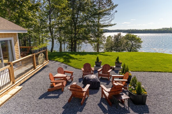 A deck with Adirondack chairs and a fire pit, overlooking a lake with trees and a house in the background.