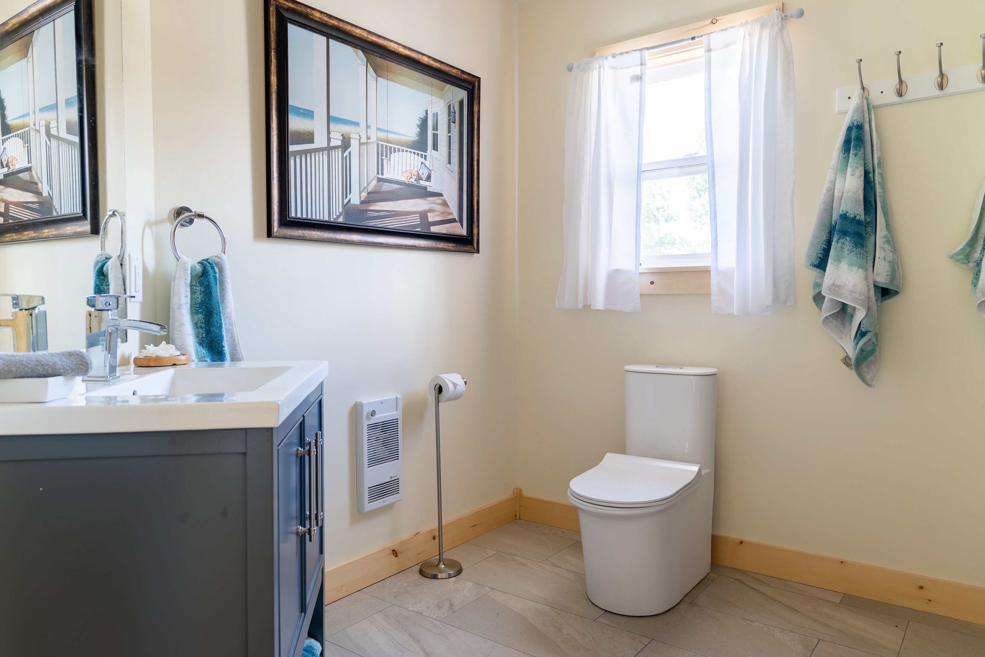 A bathroom with a toilet, sink, and a window with curtains. There are towels hanging on a rack and a mirror on the wall.