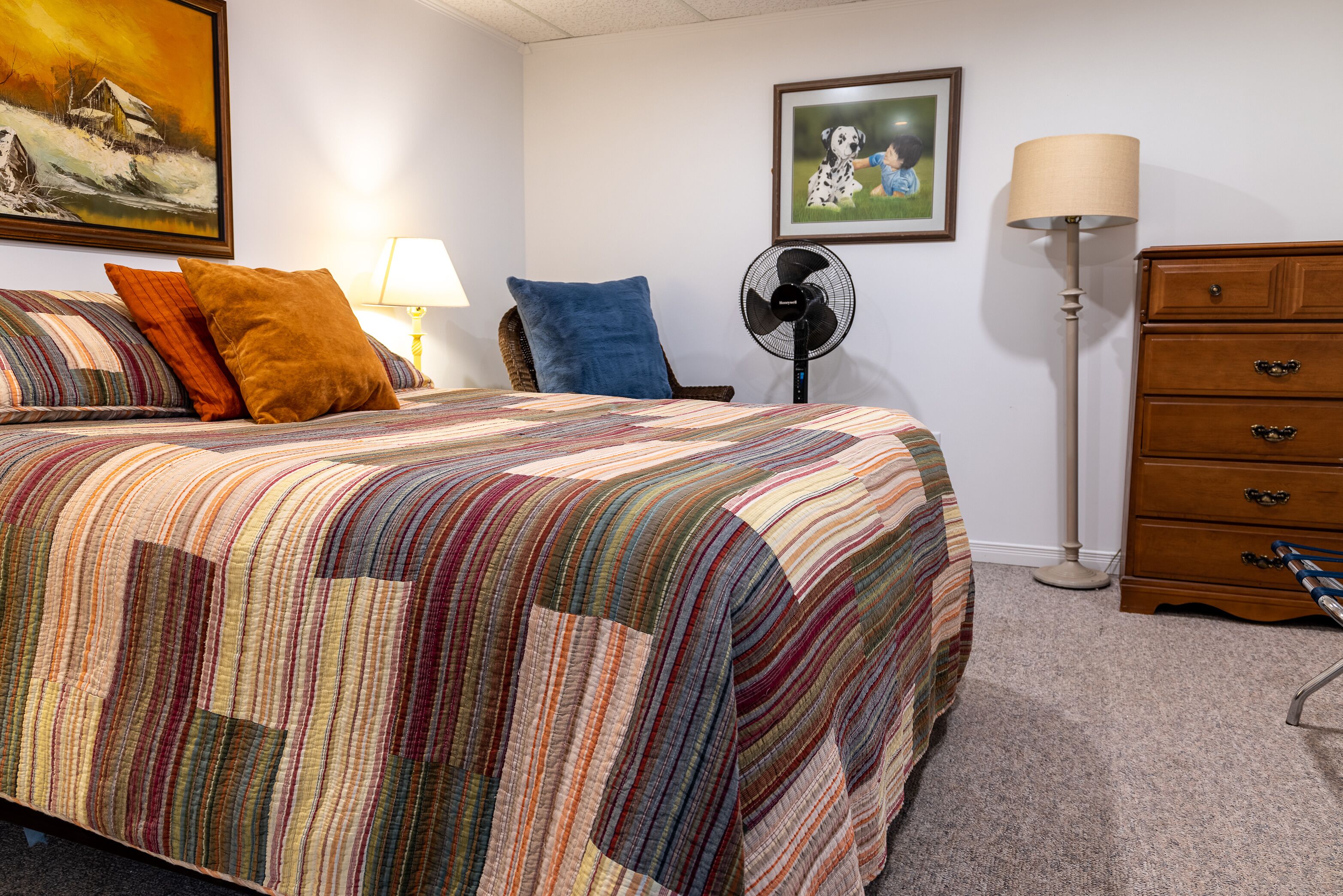 Ein Schlafzimmer mit einem großen Bett, einer Holzkommode, einem Standventilator und zwei gerahmten Bildern an der Wand.