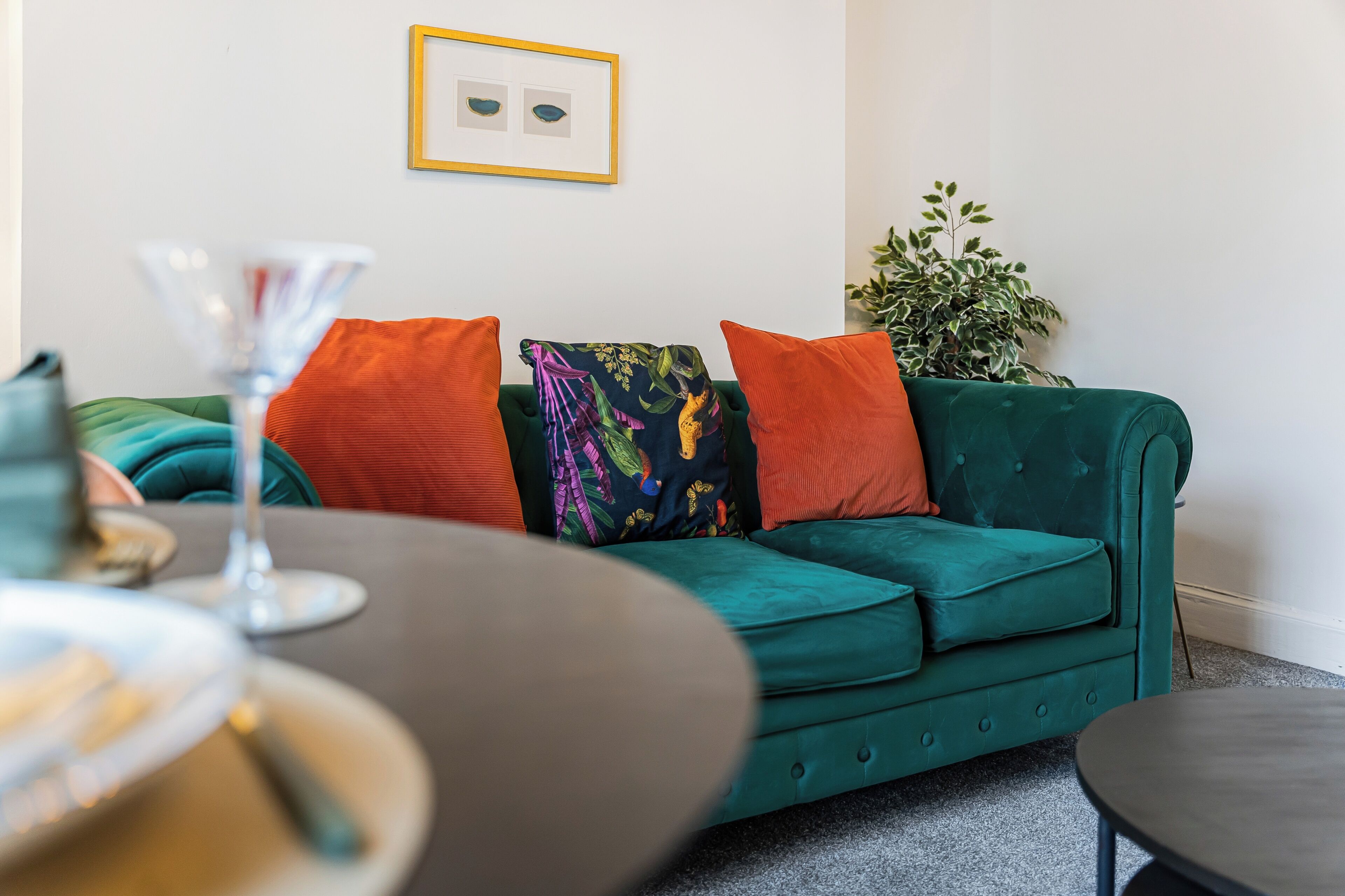 A green sofa with orange and multicolored pillows, a small table with a glass, and a framed picture on the wall.