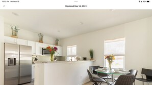 Una cocina moderna con armarios blancos, un refrigerador de acero inoxidable y un comedor con mesa y sillas de vidrio.