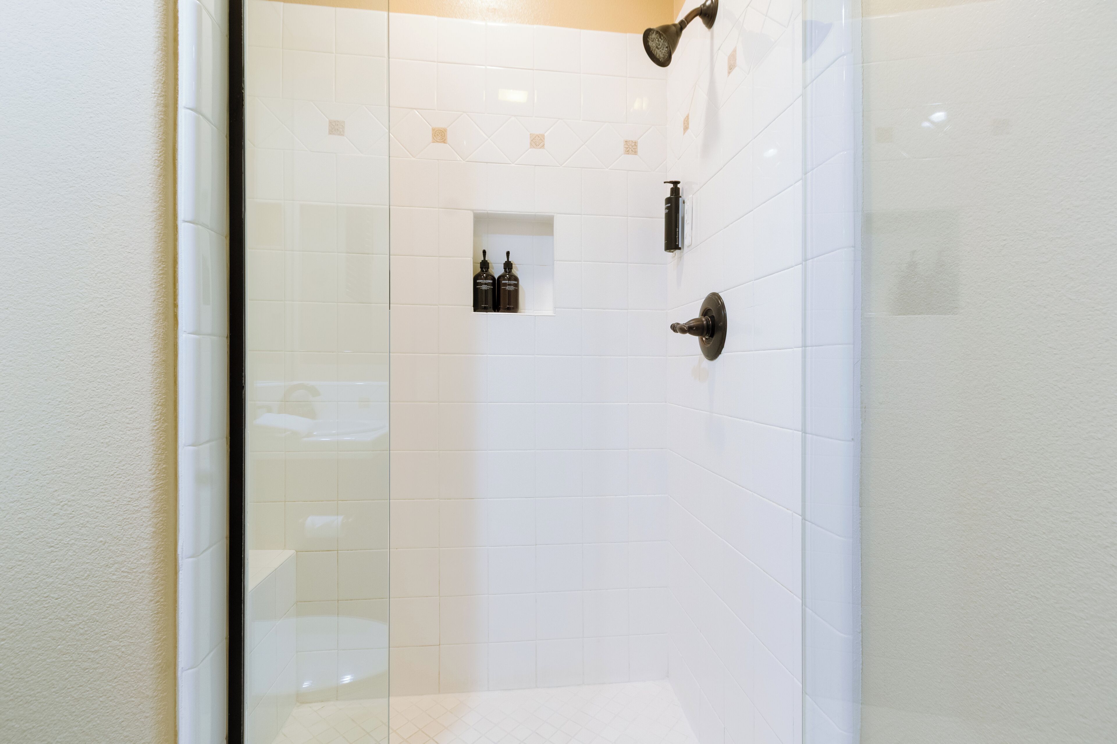 Une salle de bain carrelée de carreaux blancs, dotée d’une douche en verre et d’une petite étagère où sont rangés les articles de toilette.