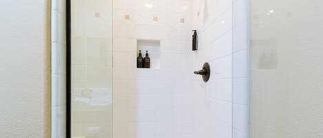 Une salle de bain carrelée de carreaux blancs, dotée d’une douche en verre et d’une petite étagère où sont rangés les articles de toilette.