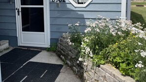 Une maison avec le numéro « 8902 » affiché sur le côté, une porte d’entrée, une petite fenêtre et un mur de pierre orné de plantes.