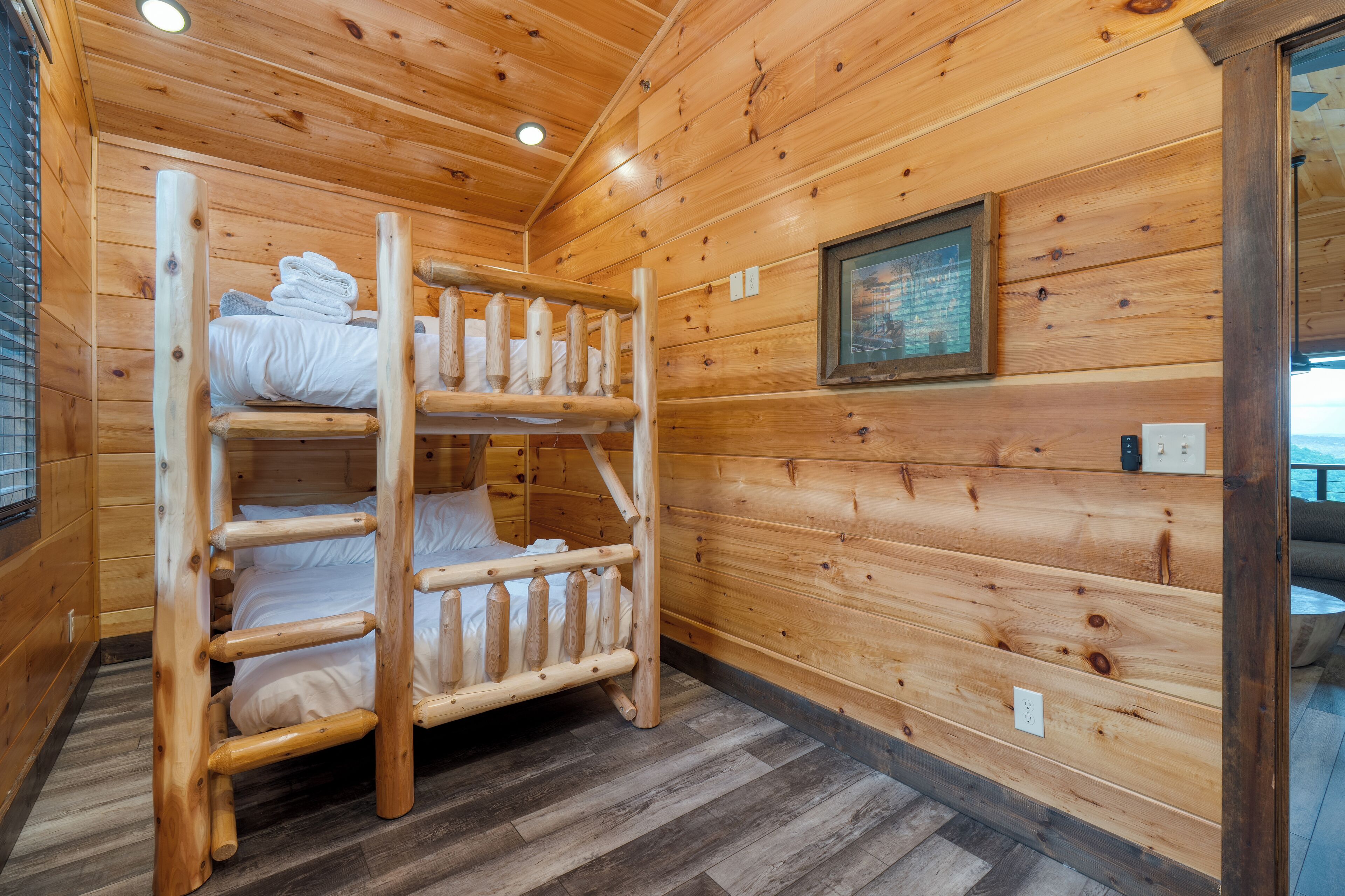 A wooden cabin with bunk beds, a framed picture on the wall, and a window with blinds.