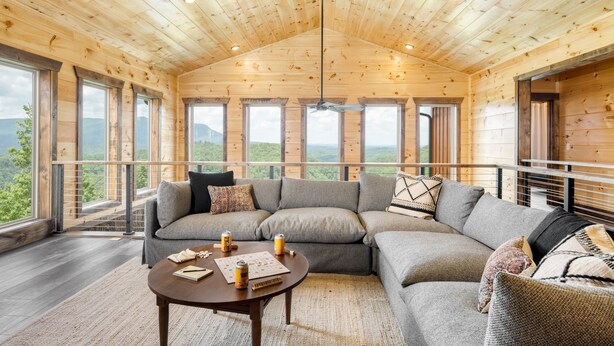 Een houten interieur met een grote hoekbank, een ronde salontafel, en een balkon met uitzicht op de bergen.