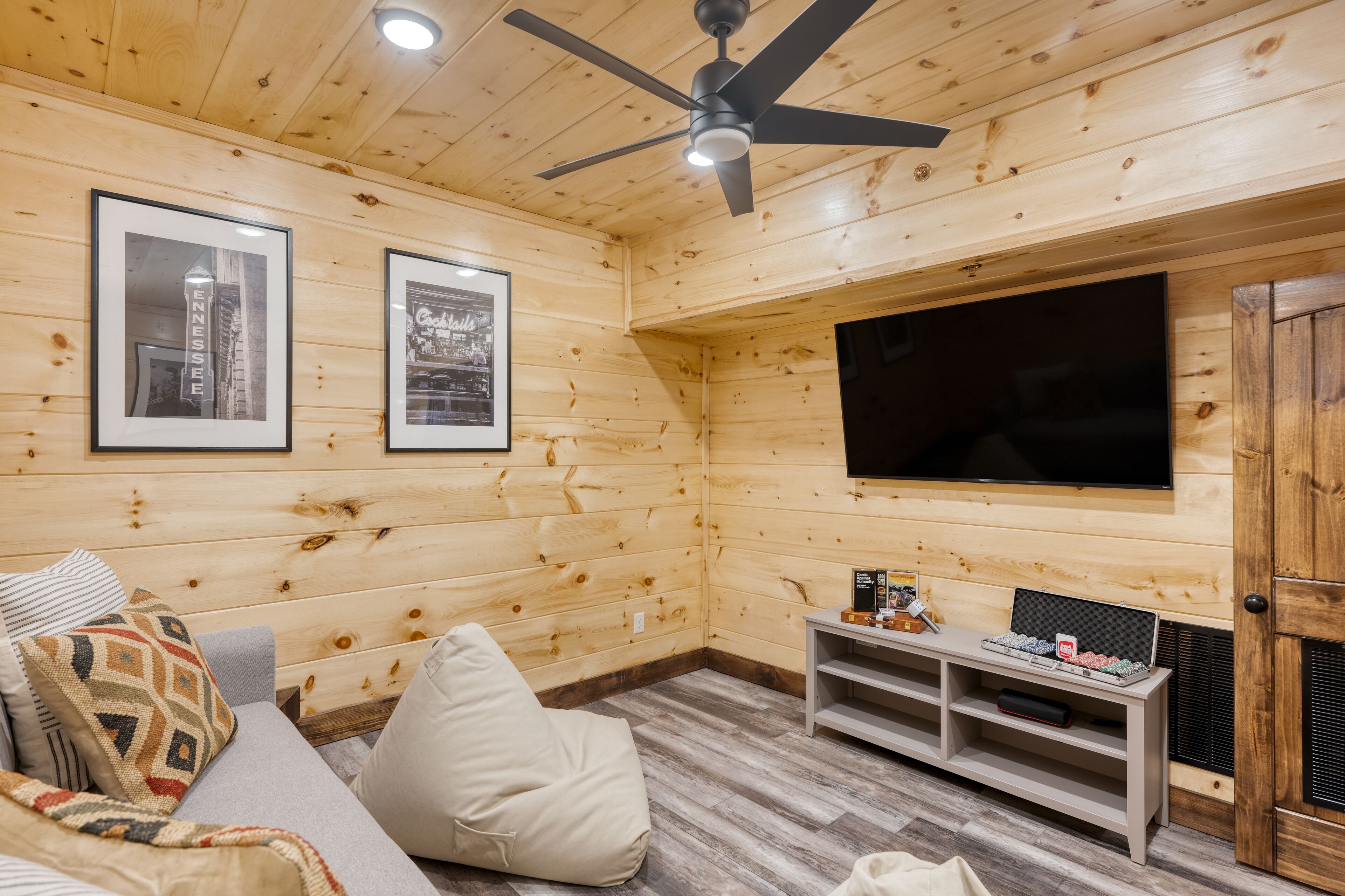 A cozy room with wooden walls, a ceiling fan, a flat-screen TV, a sofa with cushions, and a small table with items.