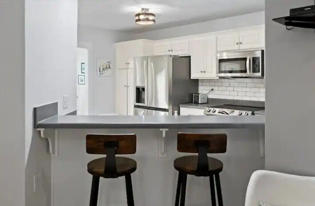 Une cuisine moderne avec des armoires blanches, un réfrigérateur en acier inoxydable, un four à micro-ondes et une cuisinière. On peut voir deux tabourets de bar au premier plan.