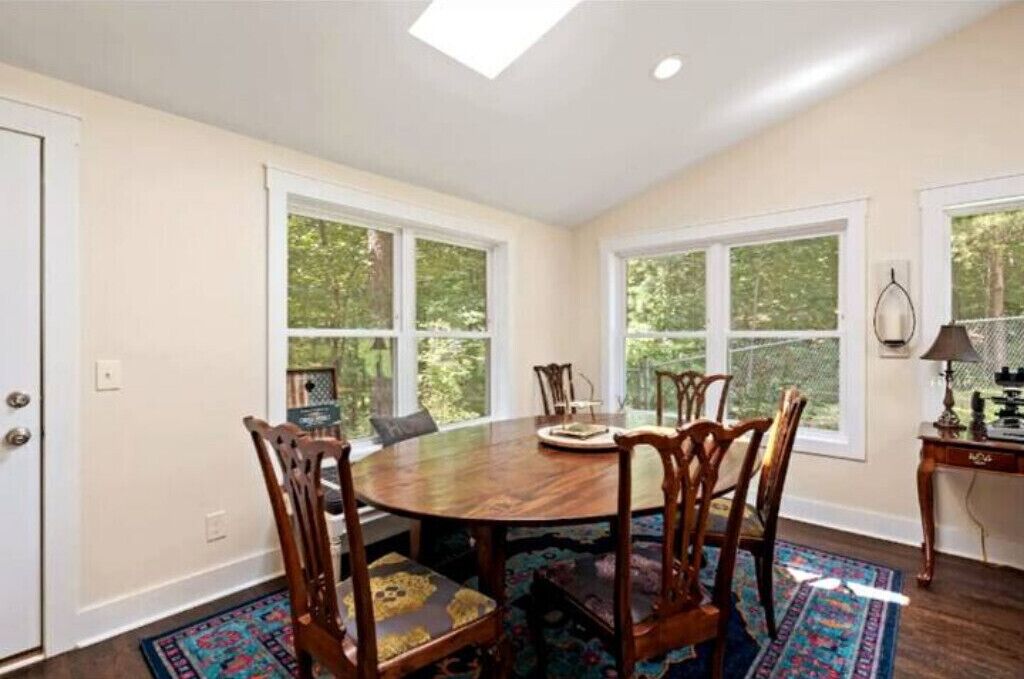 A dining room with a wooden table and chairs, large windows, and a view of greenery outside.