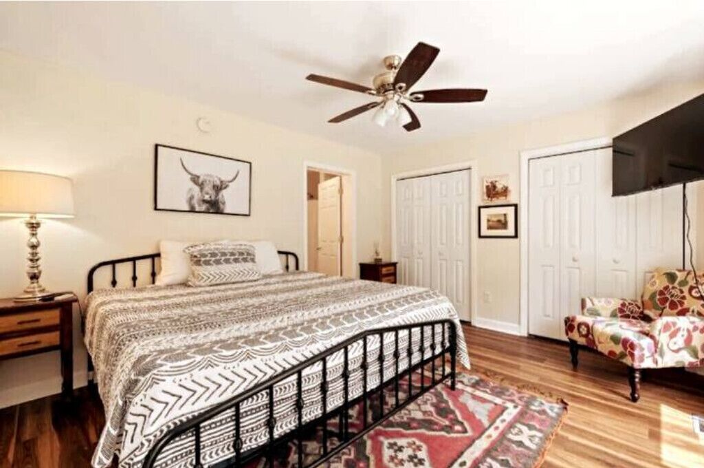 Un dormitorio con cama, ventilador de techo, mesita de noche, lámpara, silla y puerta.