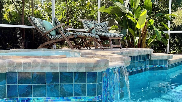 A swimming pool with tiled edges and a waterfall feature, surrounded by lounge chairs and tropical plants. - Private Heated Pool and Hottub , Pet Friendly, New Golf Cart , Close to shopping (Lady Lake)