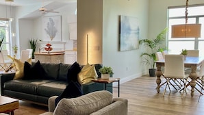 A spacious living room with a dark green sofa, a light-colored armchair, and a wooden dining table with chairs. There are ceiling fans, a chandelier, and artwork on the walls. - Private Heated Pool and Hottub , Pet Friendly, New Golf Cart , Close to shopping (Lady Lake)