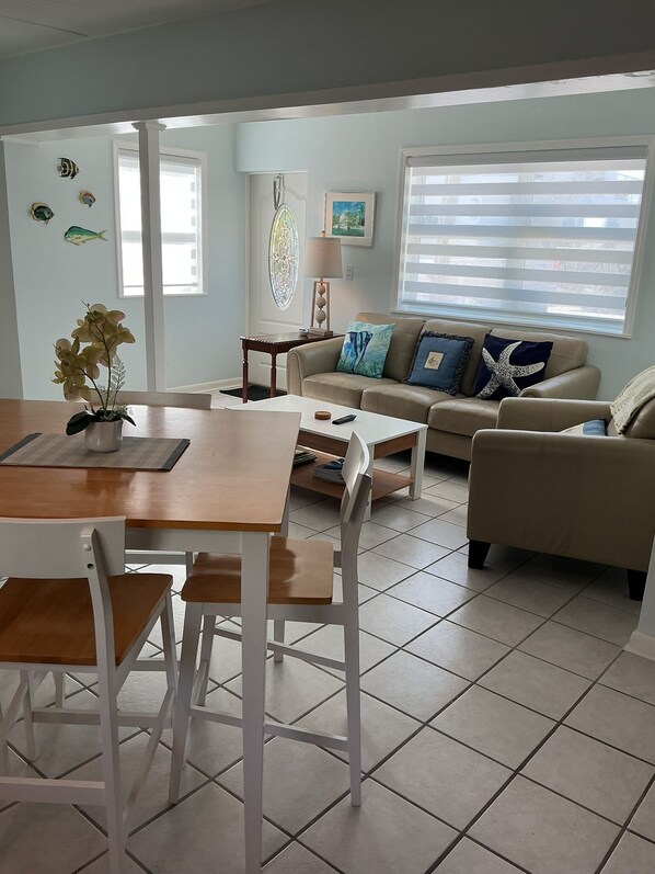 A living room with a dining table, chairs, a sofa, and a coffee table. There are decorative fish on the wall, a lamp, and a window with blinds.