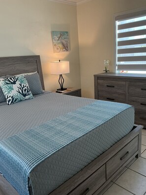 A neatly made bed with a quilted cover, a bedside lamp, a nightstand, a wooden dresser, and a window with blinds. - Private Pool & Dock (Marathon)