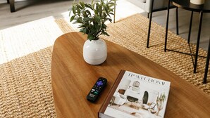 Een woonkamer met een houten salontafel, een afstandsbediening en een tijdschrift. Er staat een televisie op een dressoir met decoratieve objecten en een plant. Ook is er een witte fauteuil en een klein tafeltje met een schaal te zien.