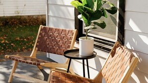 Twee houten stoelen met gevlochten zitting en rugleuning, een klein rond tafeltje en een potplant op een betonnen terras voor een wit gebouw met een raam.