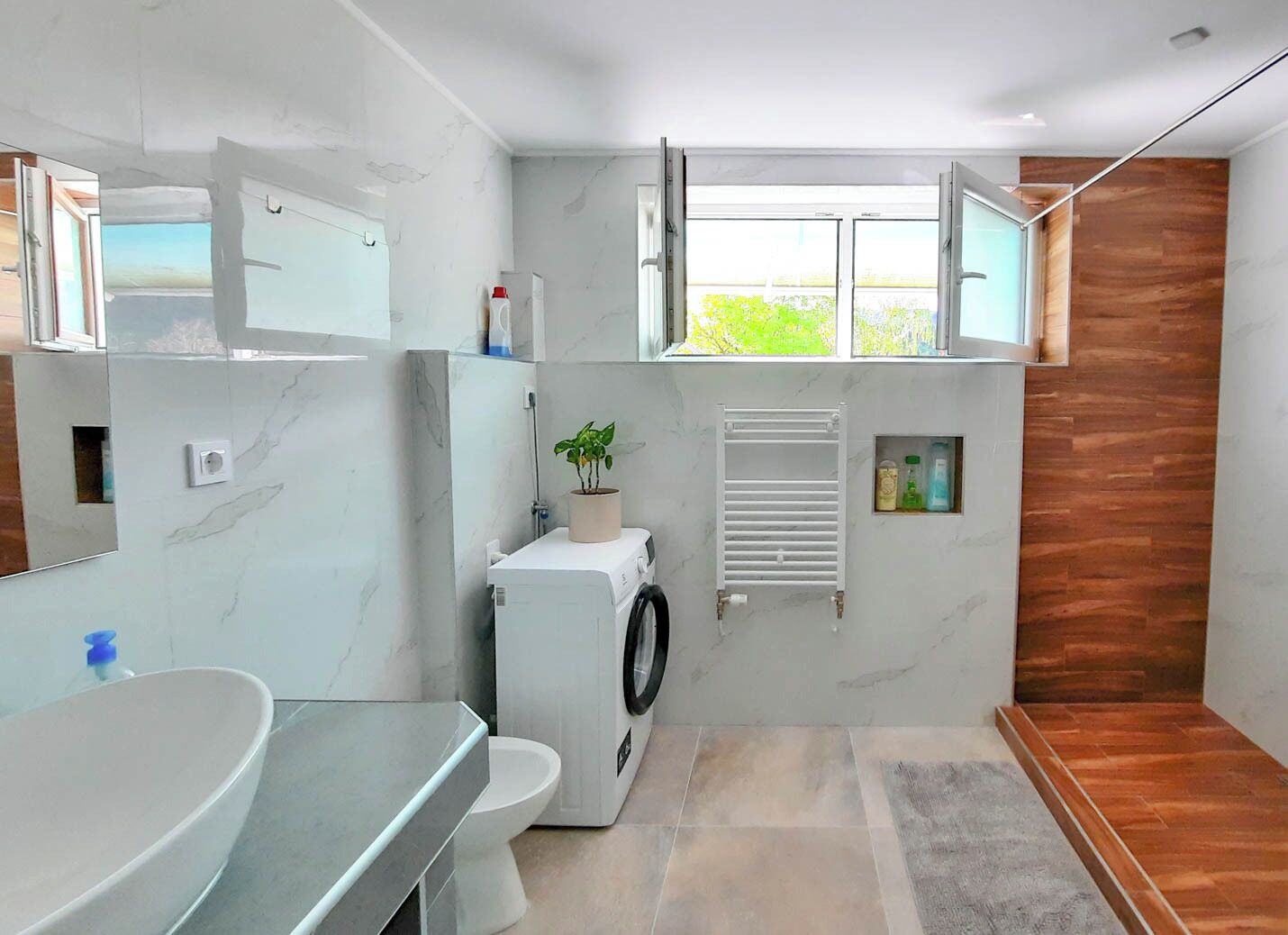 Une salle de bain moderne avec un grand miroir, un lavabo, des toilettes, un lave-linge et une fenêtre donnant sur un espace verdoyant.