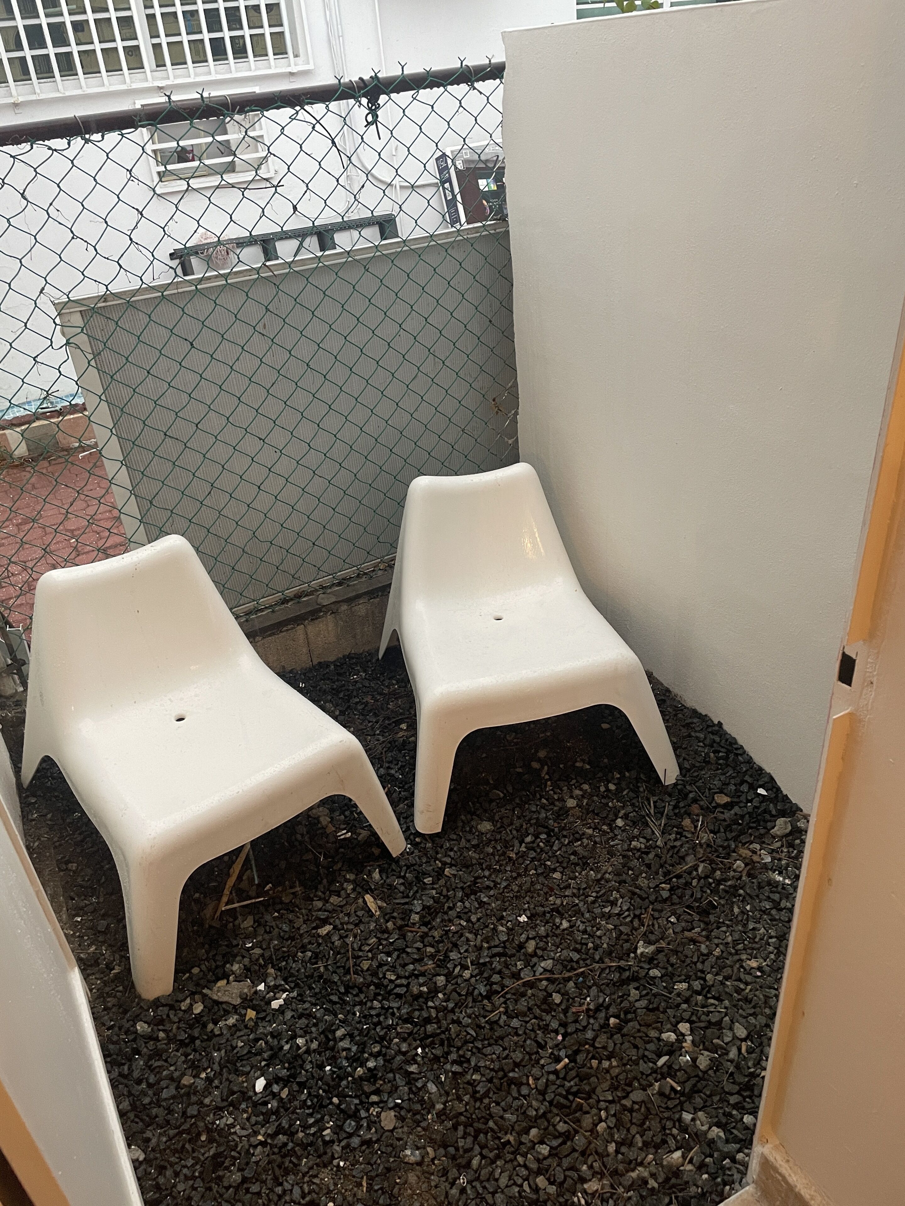 Dos sillas blancas de plástico en una esquina, con una cerca de malla metálica y una pared blanca al fondo.