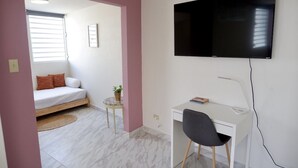 A compact room with a bed, a desk, a chair, a TV mounted on the wall, and a small table with a plant. - The Emerald (San Juan)