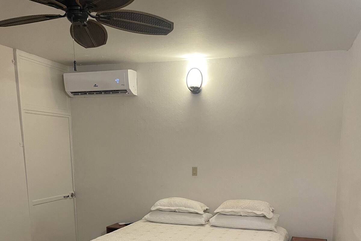 Quarto com cama, mesa de cabeceira, banquinho, ventoinha no teto, ar-condicionado e luminária na parede.