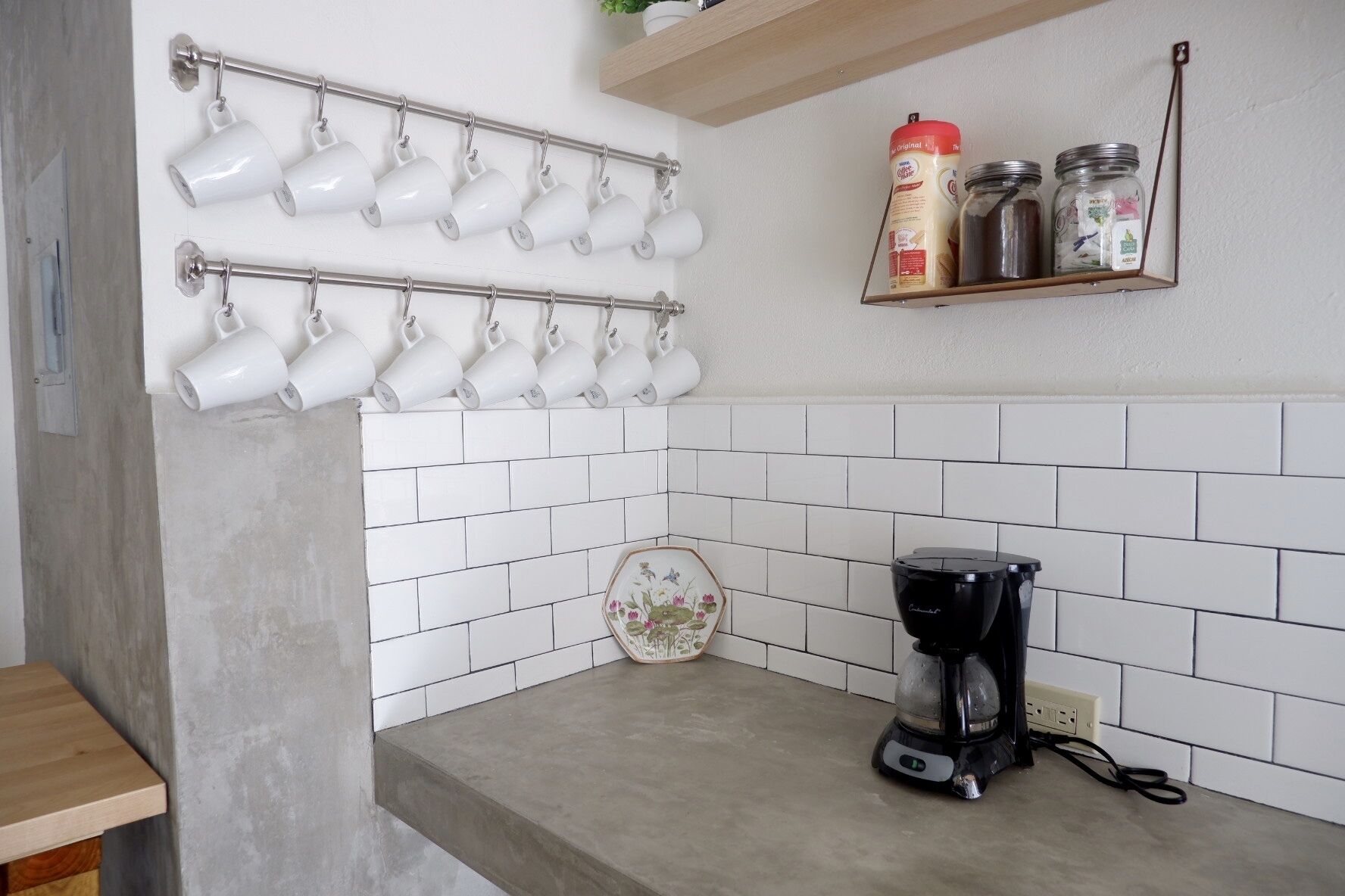 Una cocina con salpicadero de azulejos blancos, una cafetera, una repisa con frascos y un escurreplatos para tazas.