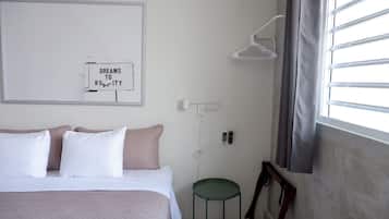 Un dormitorio con cama, almohadas, una mesita de noche, una lámpara de pared, una ventana con persianas y un cuadro en la pared.