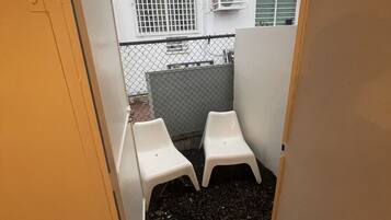 Un couloir étroit mène à un petit espace extérieur où se trouvent deux chaises en plastique blanc et une clôture en métal.