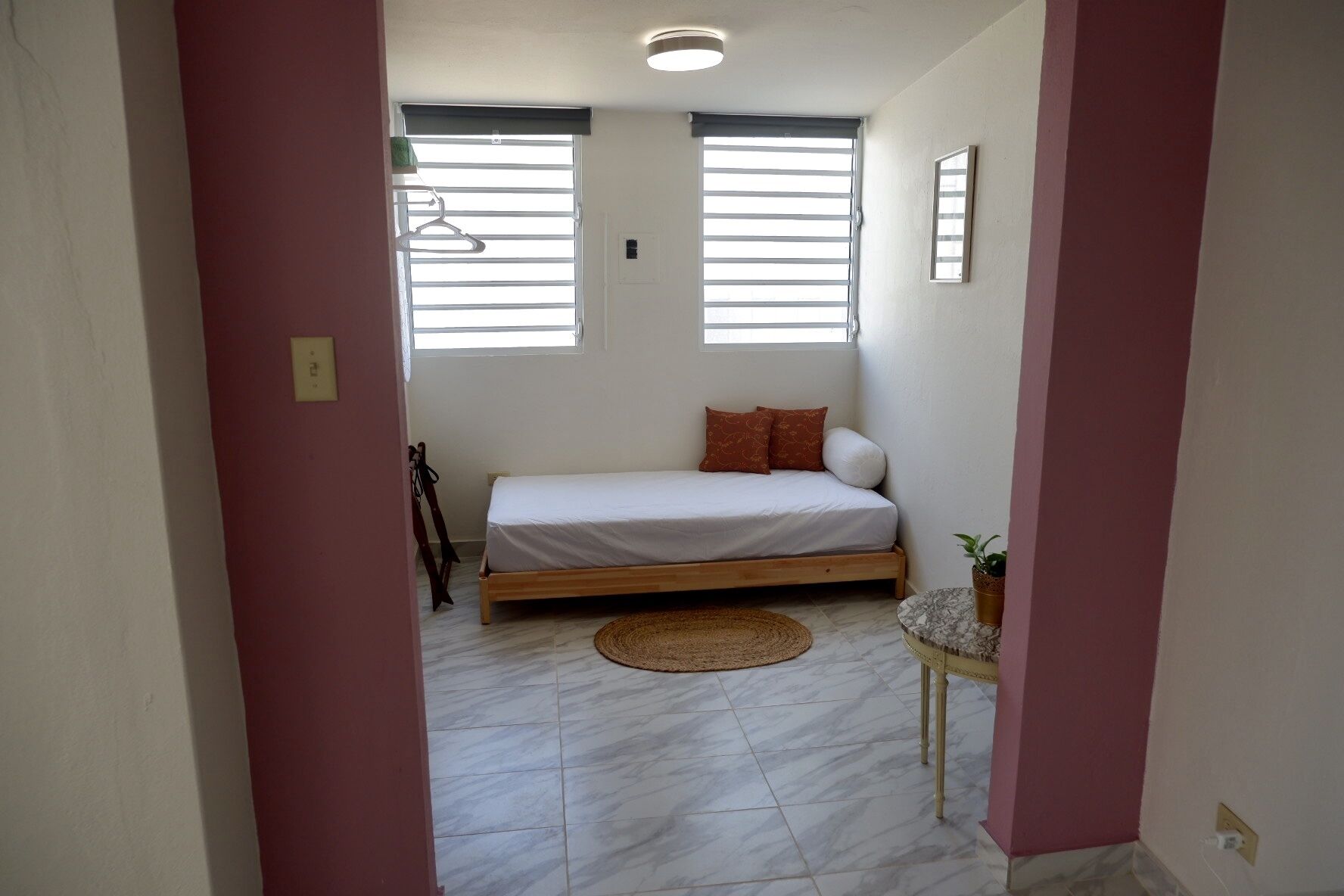 A bedroom with a bed, a small table, a potted plant, and a window with blinds.