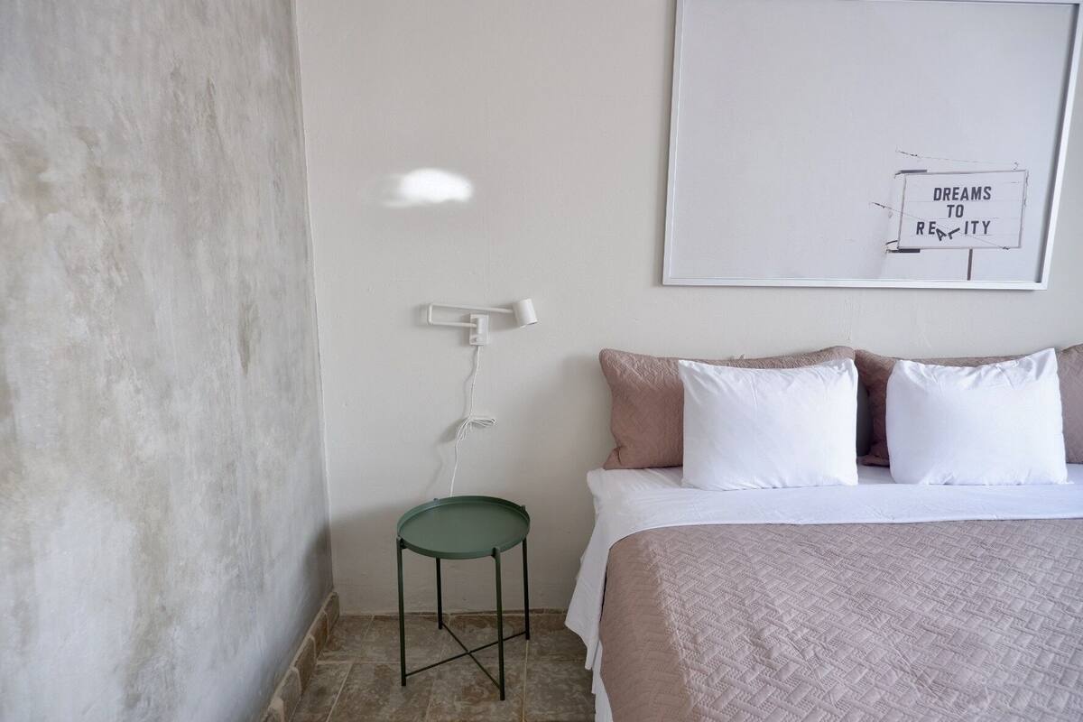 Quarto com cama, almofadas, uma cadeira verde, parede branca e um letreiro emoldurado.
