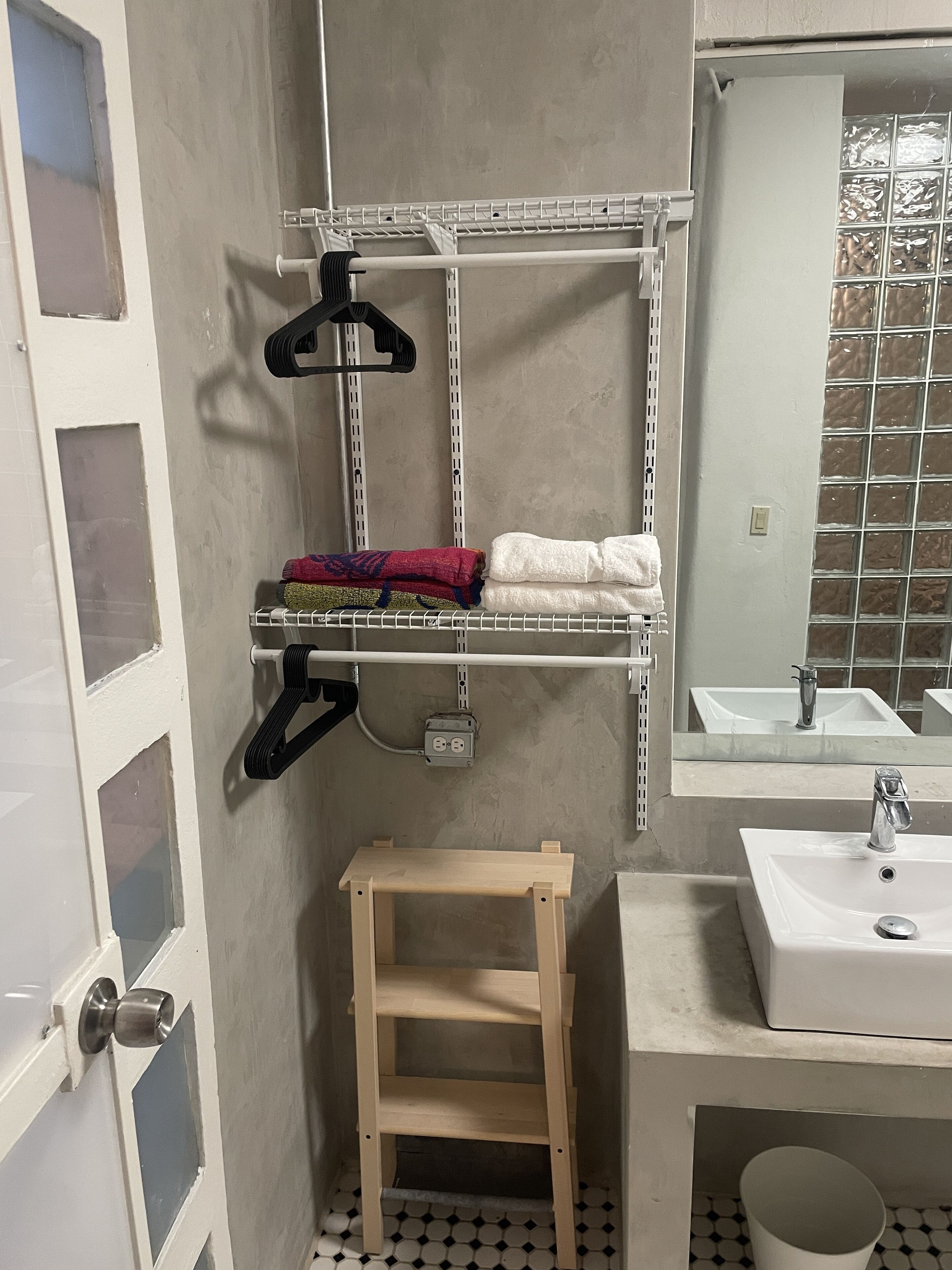 Un baño con un lavamanos blanco, un espejo, un perchero para toallas y un taburete de madera.