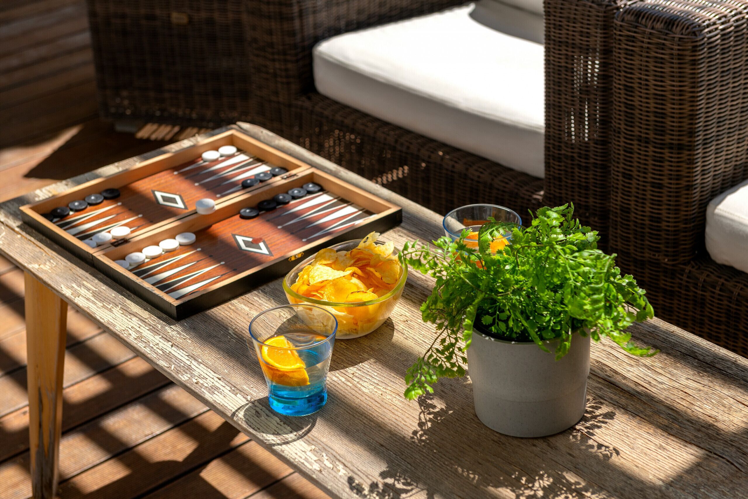 Ein Holztisch mit einem Brettspiel, einer Snackschale, einem Glas mit Getränk und einer Topfpflanze.