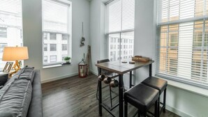 A modern, well-lit room with a dining table, two chairs, a sofa, and large windows with blinds. - 725- Big 2 bedroom/king beds/Corner Unit/ + parking (Milwaukee)