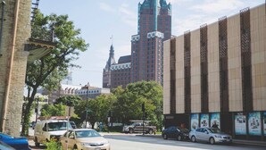 Straßenansicht mit einem hohen Gebäude im Hintergrund und einem großräumigen Gebäude mit vertikalen Säulen auf der rechten Seite. Entlang der Straße sind parkende Autos zu sehen.