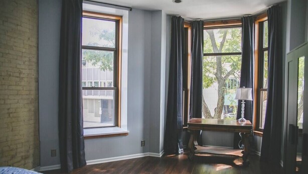 Ein Schlafzimmer mit großem Fenster, einem Holztisch und einem Bett mit gemusterten Bettbezügen.
