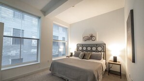 A bedroom with a large window, a bed with a headboard, a bedside table, and a wall-mounted artwork. - 325- Big Corner condo w/2 king bedrooms/Free parking (Milwaukee)