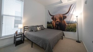 A bedroom with a bed, a nightstand, a lamp, a large wall tapestry of a bull, and a window with blinds.