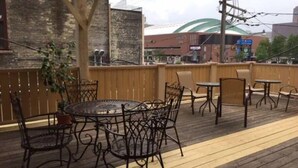 Un patio al aire libre con una mesa redonda y sillas, cubierta de madera y vistas a edificios al fondo.