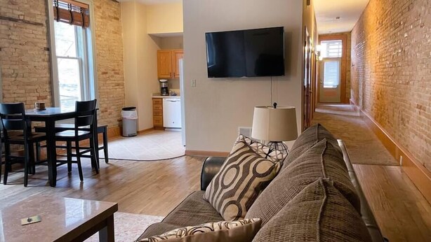 Una sala de estar con un sofá, una mesa de comedor y sillas. Se observa una cocina al fondo con armarios y un televisor montado en la pared.
