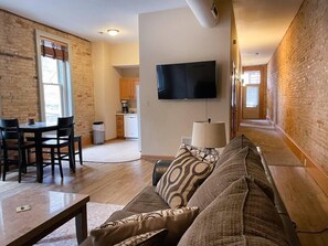 A living room with a sofa, a dining table, and chairs. There is a kitchen in the background with cabinets and a television mounted on the wall. - Unit B- Large Condo w/Patio/ Old World 3rd+Fiserv (Milwaukee)