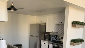 A modern kitchen with white cabinets, a marble countertop, a stainless steel refrigerator, a microwave, and an oven. There are two bar stools and a hanging light fixture.