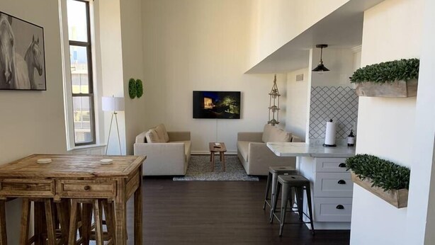 A modern living room with a wooden dining table, two sofas, a TV, and a kitchen area with white cabinets and a hanging plant.